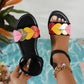 Women's black flat sandals adorned with colorful heart embellishments in pink, yellow, red, and gold, featuring a black elastic ankle strap and white contrast stitching, worn on a foot against a blue patterned background.