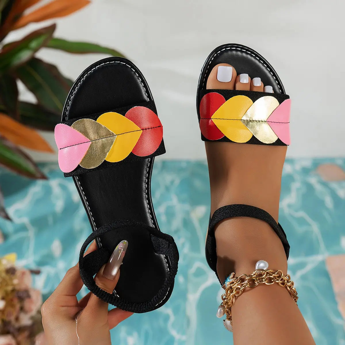 Women's black flat sandals adorned with colorful heart embellishments in pink, yellow, red, and gold, featuring a black elastic ankle strap and white contrast stitching, worn on a foot against a blue patterned background.