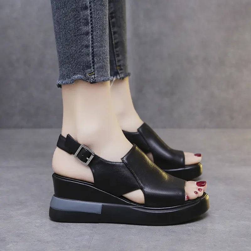 Black Kaelos wedge sandals with a crossover peep-toe, adjustable slingback strap, and a grey accent stripe on the sole, shown from a three-quarter angle.