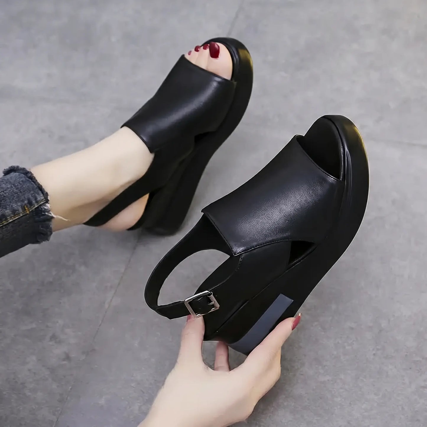A pair of Kaelos black wedge sandals with a crossover peep-toe design and adjustable slingback strap, shown on a foot with distressed denim and held by a hand. Features a grey accent stripe on the wedge.