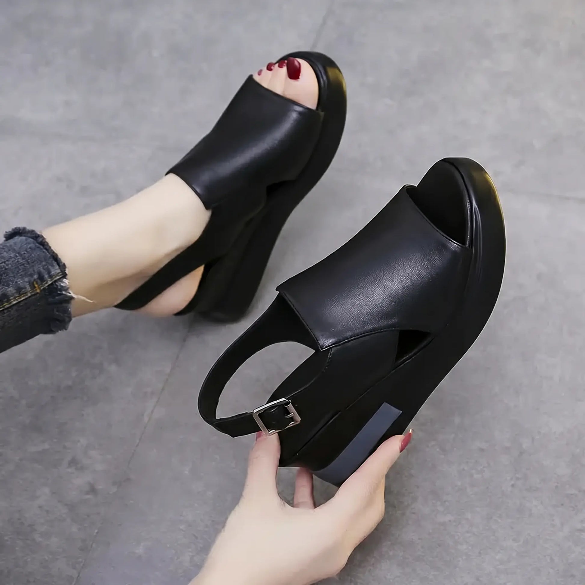 A pair of Kaelos black wedge sandals with a crossover peep-toe design and adjustable slingback strap, shown on a foot with distressed denim and held by a hand. Features a grey accent stripe on the wedge.