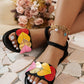 Kaelos flat sandals adorned with vibrant multi-colored heart embellishments, featuring a black elasticated ankle strap and a decorative charm bracelet, styled on feet with a summery backdrop of a straw hat and flowers.
