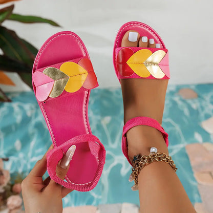 Close-up of vibrant pink Kaelos flat sandals adorned with multi-colored heart embellishments and a black elasticated ankle strap, styled on a model's foot with a blurred water background.