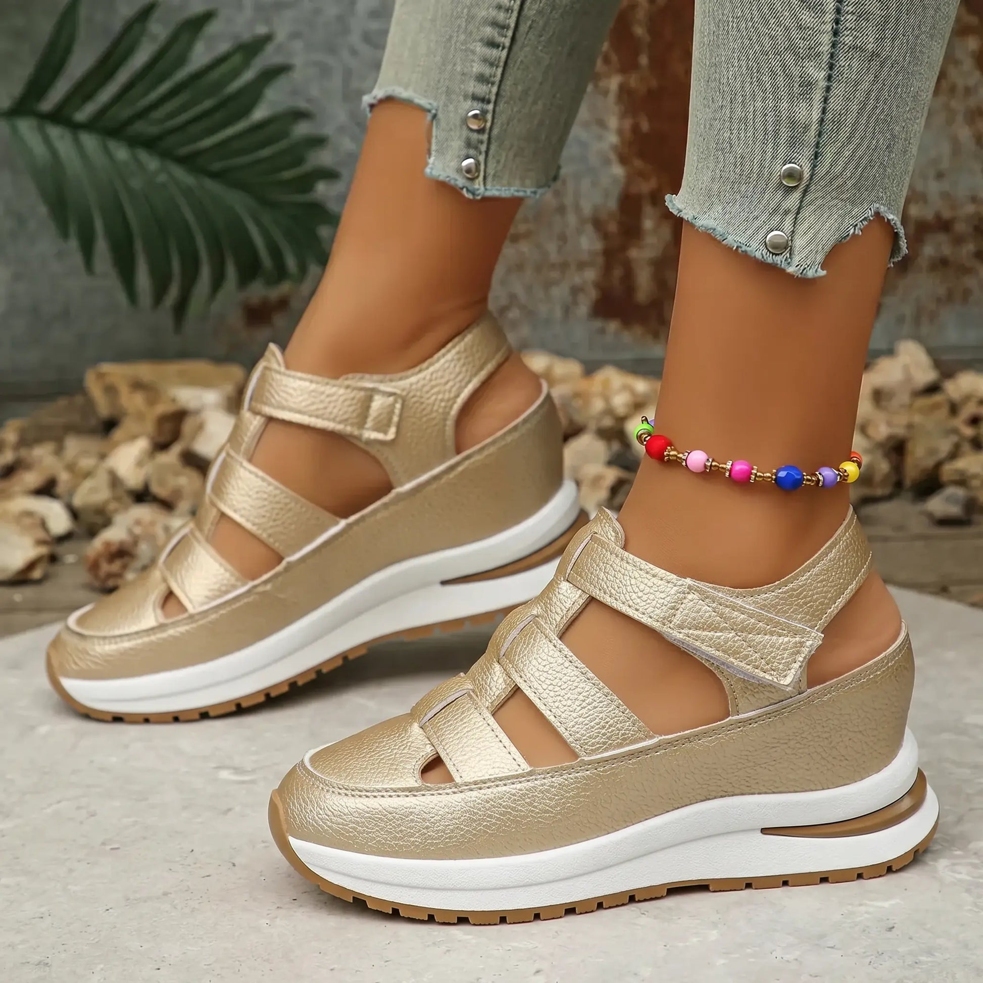 Kaelum women's subtle gold metallic platform wedge sandals with cut-out straps are shown on a model's feet with cropped jeans and a colorful anklet, on a textured backdrop.
