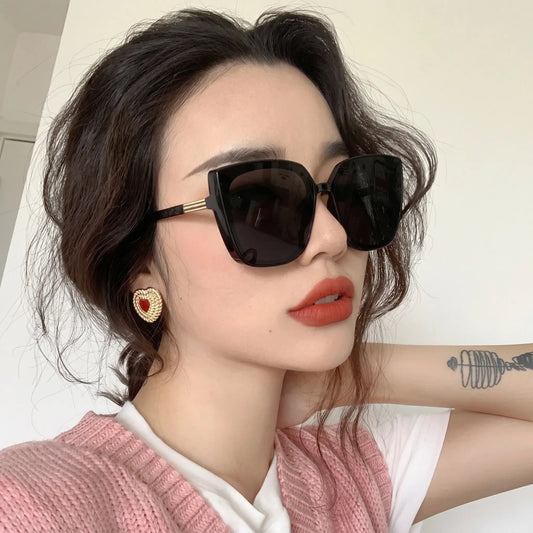 Elegant woman wearing Kaelum oversized angular butterfly sunglasses with glossy black frames, grey lenses, and gold temple accents, styled for a chic look.