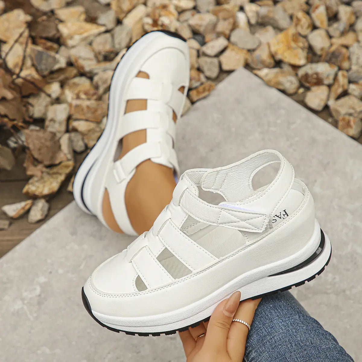 A pair of white Kaelum platform wedge sandals with cut-out straps, shown on a foot and held by a hand against a textured background.