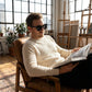 Man wearing KAEON oversized square sunglasses in black, sitting in a leather chair reading a book in a sunlit loft studio.