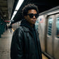 KAEON oversized square sunglasses black worn by a man in a leather jacket on a subway platform, showing the bold architectural frame.