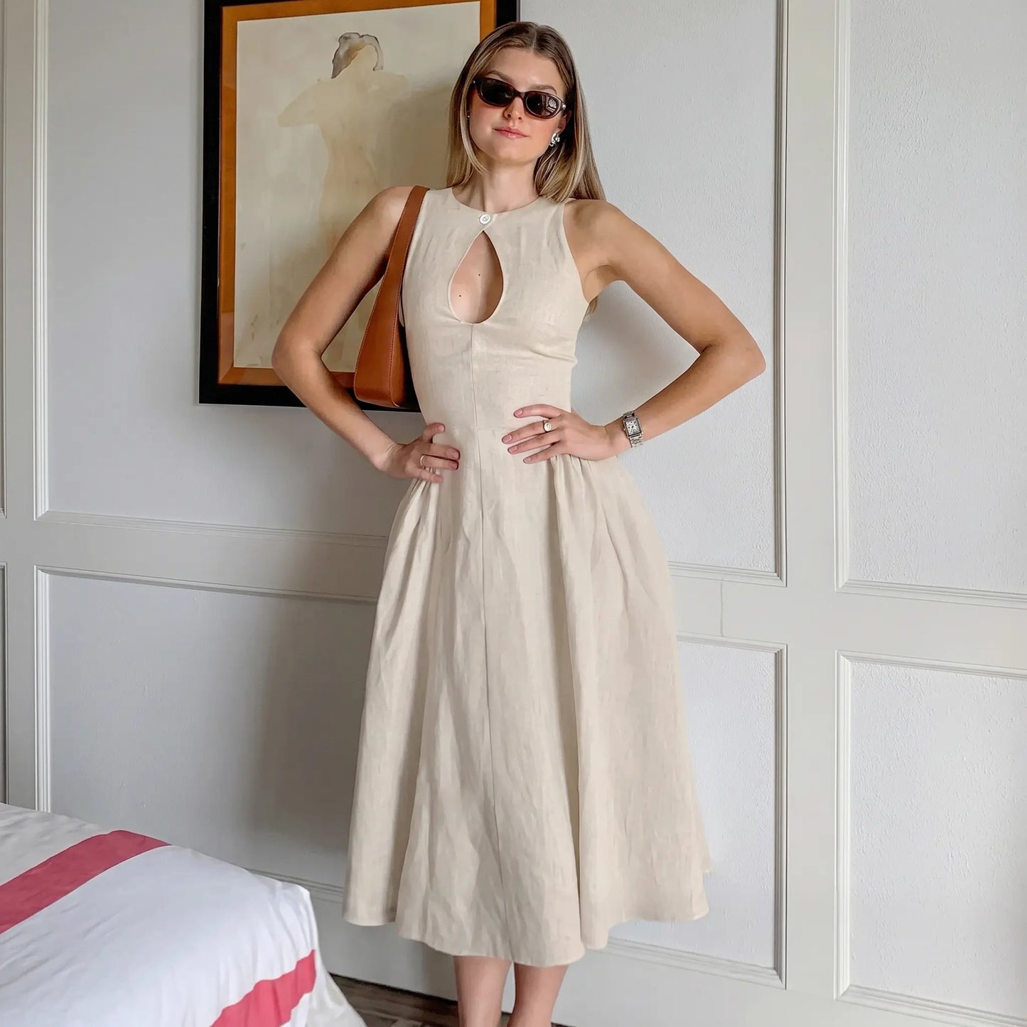 A woman models a sleeveless, natural beige linen-blend midi dress with a keyhole neckline and gathered A-line skirt, styled with sunglasses and a handbag.