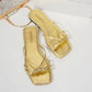 Metallic gold flat sandals with a square toe and delicate knotted straps, displayed on a white studio surface with a clutch bag. These Kallia summer slip-ons offer chic comfort.