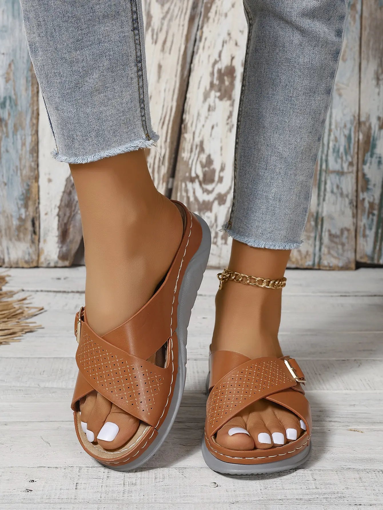 Kalyx women's brown faux leather wedge sandals featuring elegant criss-cross straps with micro-perforations and a gold-tone adjustable buckle, styled with denim jeans on a wooden floor.