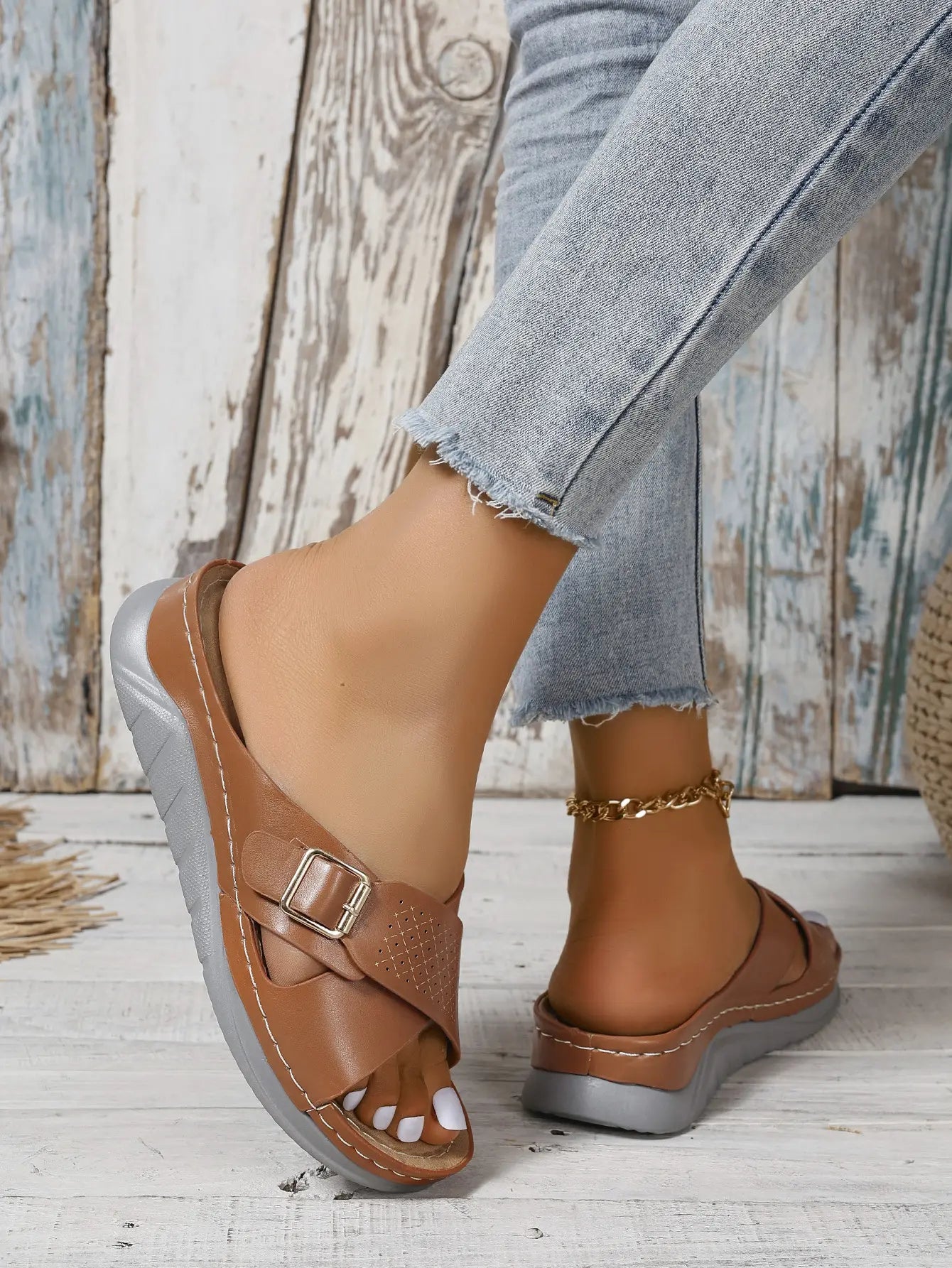 Kalyx women's brown faux leather wedge sandals with elegant criss-cross straps, gold-tone buckle, and micro-perforations, shown worn with denim jeans.