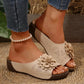 Beige women's slide sandals featuring a prominent 3D fabric flower embellishment on the vamp, open toe, low wedge platform sole, and perforated faux leather detailing, shown with jeans and a pearl anklet.