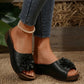 Black Kavlo women's open-toe slide sandals featuring a prominent 3D fabric flower embellishment on the vamp and a low wedge platform sole, shown in a lifestyle shot with jeans and a pearl anklet.