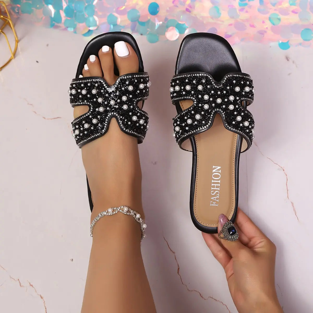 Elegant black Kezia flat sandals adorned with lustrous pearls and sparkling rhinestones, worn on a foot with white toenails and a delicate anklet.