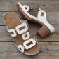 White Khoros heeled slide sandals featuring a geometric gold-tone hardware strap and a chunky block heel with a textured brown platform. View of a pair of women's stylish mule sandals on a wooden surface.