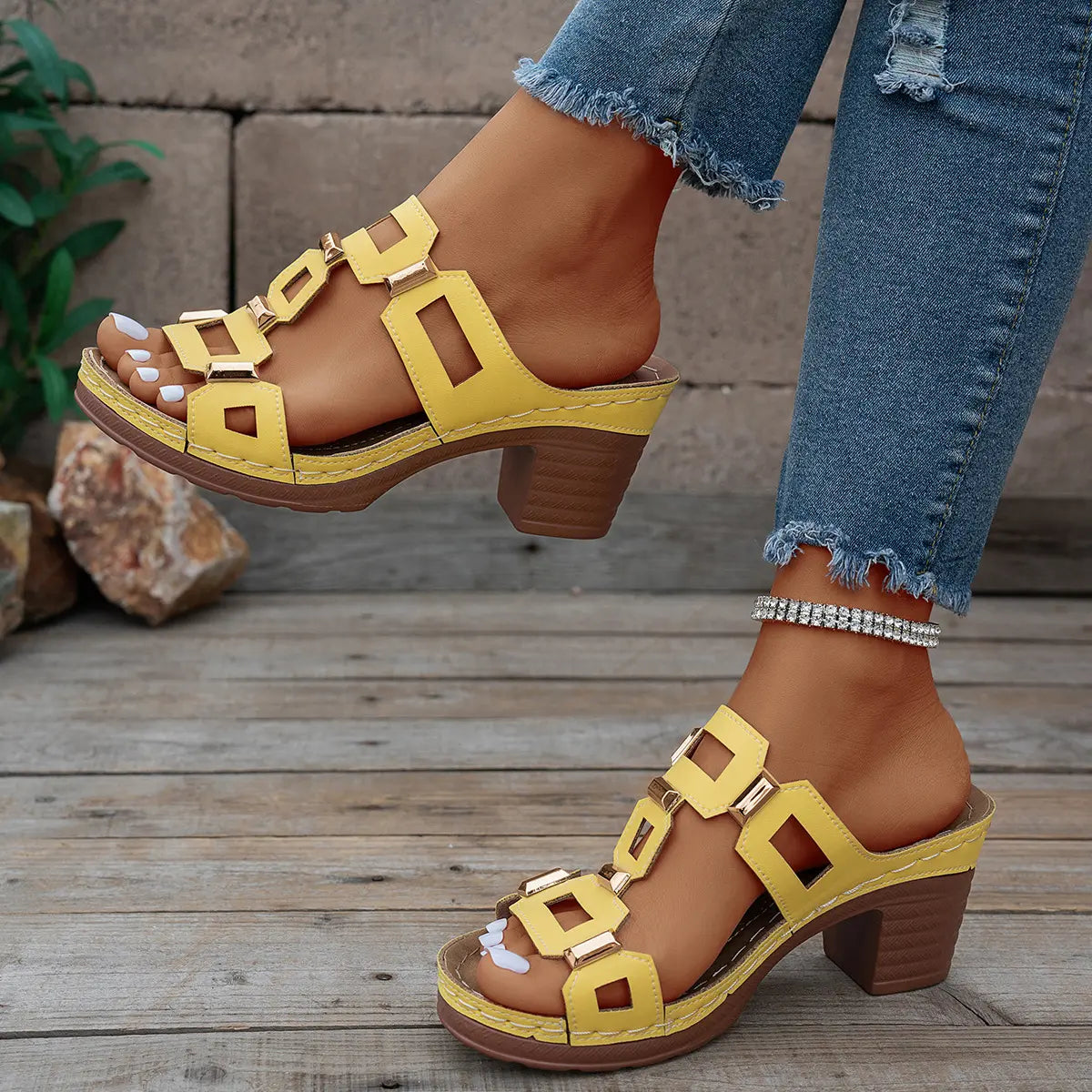 Woman's yellow heeled platform slide sandals with geometric gold hardware, styled with distressed denim jeans and a silver anklet, shown on a rustic wooden surface.