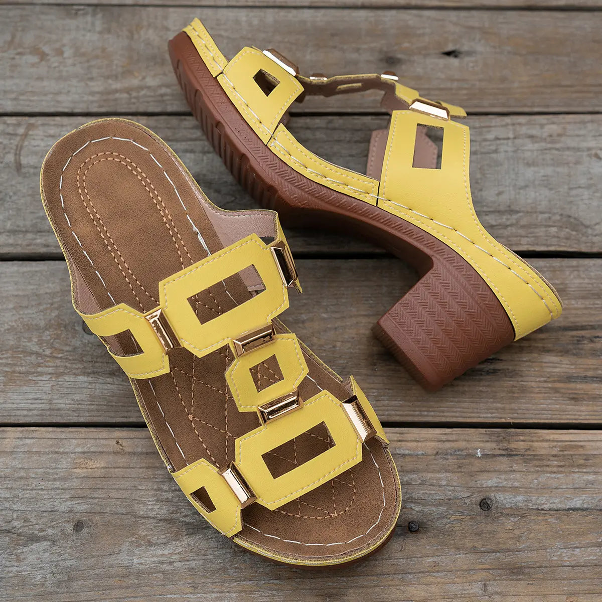 Yellow Khoros heeled platform slide sandals featuring geometric gold-tone hardware and a chunky block heel, displayed on a wooden surface.