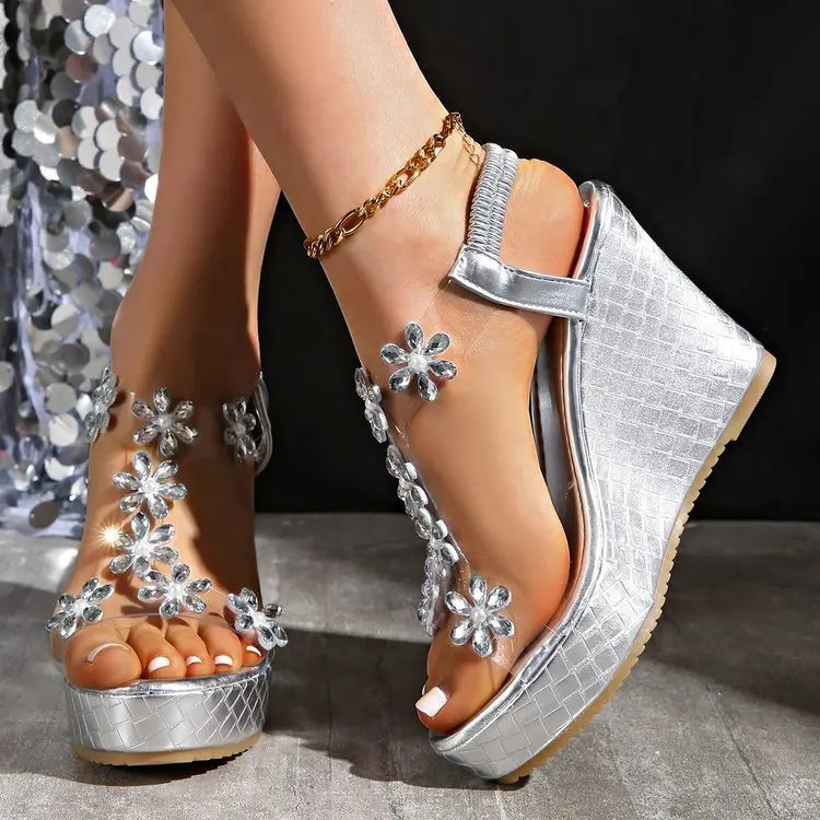 Women's silver wedge sandals adorned with sparkling crystal floral embellishments on clear transparent straps, featuring a textured metallic wedge heel and platform sole, worn with an anklet.