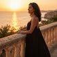 Model wearing a black Khozun open-back maxi dress with thin spaghetti straps, admiring a golden sunset over the ocean from a stone balcony.