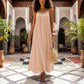 Khozun Bohemian Open-Back Maxi Dress in soft apricot, worn by a model walking through an ornate Moroccan courtyard with potted plants and patterned tiles. Features delicate spaghetti straps and a gathered scoop neckline.