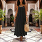 Woman in a Khozun Midnight Black Bohemian maxi dress featuring a voluminous tent silhouette, delicate spaghetti straps, and gathered scoop neckline, walking through an elegant tiled courtyard.