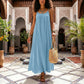 A woman wears a flowing sky blue Khozun Bohemian Open-Back Maxi Dress with thin spaghetti straps and a gathered scoop neckline, walking through an ornate tiled courtyard. The voluminous, floor-length tent silhouette drapes gracefully as she carries a woven bag.