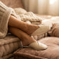 Alabaster Beige vegan leather V-vamp mule flats worn by a person resting on cozy, soft cushions, highlighting the shoes' elegant and comfortable slip-on style.
