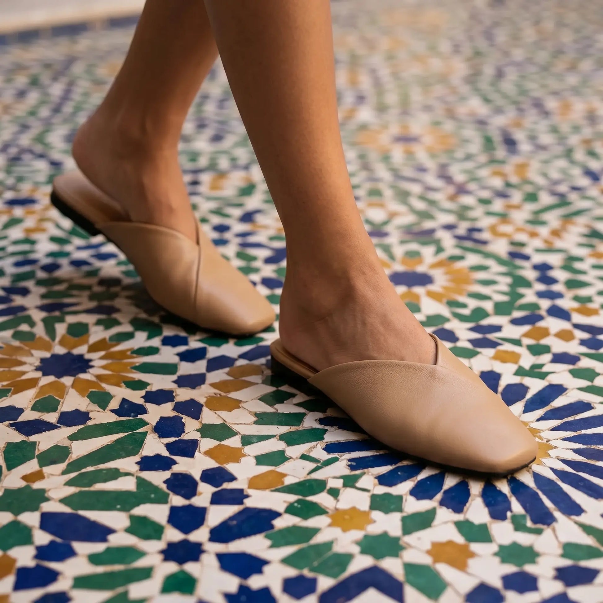 Khyren Alabaster Beige vegan leather V-vamp mule flats with square toes, worn by a model walking on a vibrant mosaic tiled floor.
