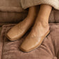 Close-up lifestyle shot of Khyren Desert Khaki vegan leather mule flats on a person's feet, featuring an asymmetrical V-cut vamp and rounded square toe, resting on a brown cushion.