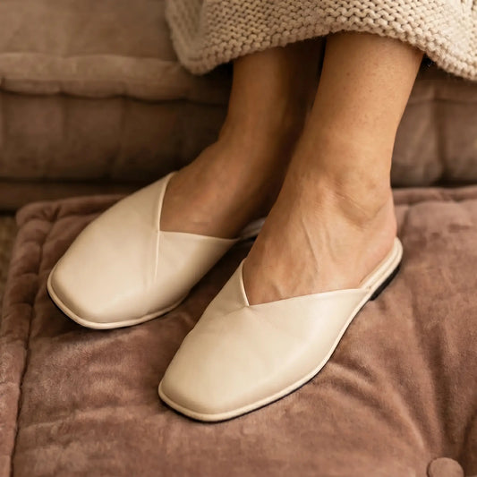 Alabaster Beige Khyren V-vamp mule flats on feet, with asymmetrical vamp and rounded square toe, resting on a soft brown cushion.