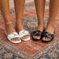Khyzal black and white square toe slide sandals with gold-tone horseshoe buckles, worn by women on a patterned rug. One foot displays a pearl anklet.