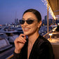Smiling woman wearing Kivago black metal cat-eye sunglasses with dark tinted lenses at a stylish outdoor restaurant overlooking a marina at dusk.