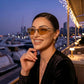Smiling woman wearing Kivago gunmetal cat-eye sunglasses with amber tinted lenses, enjoying an evening at a marina with city lights in the background.