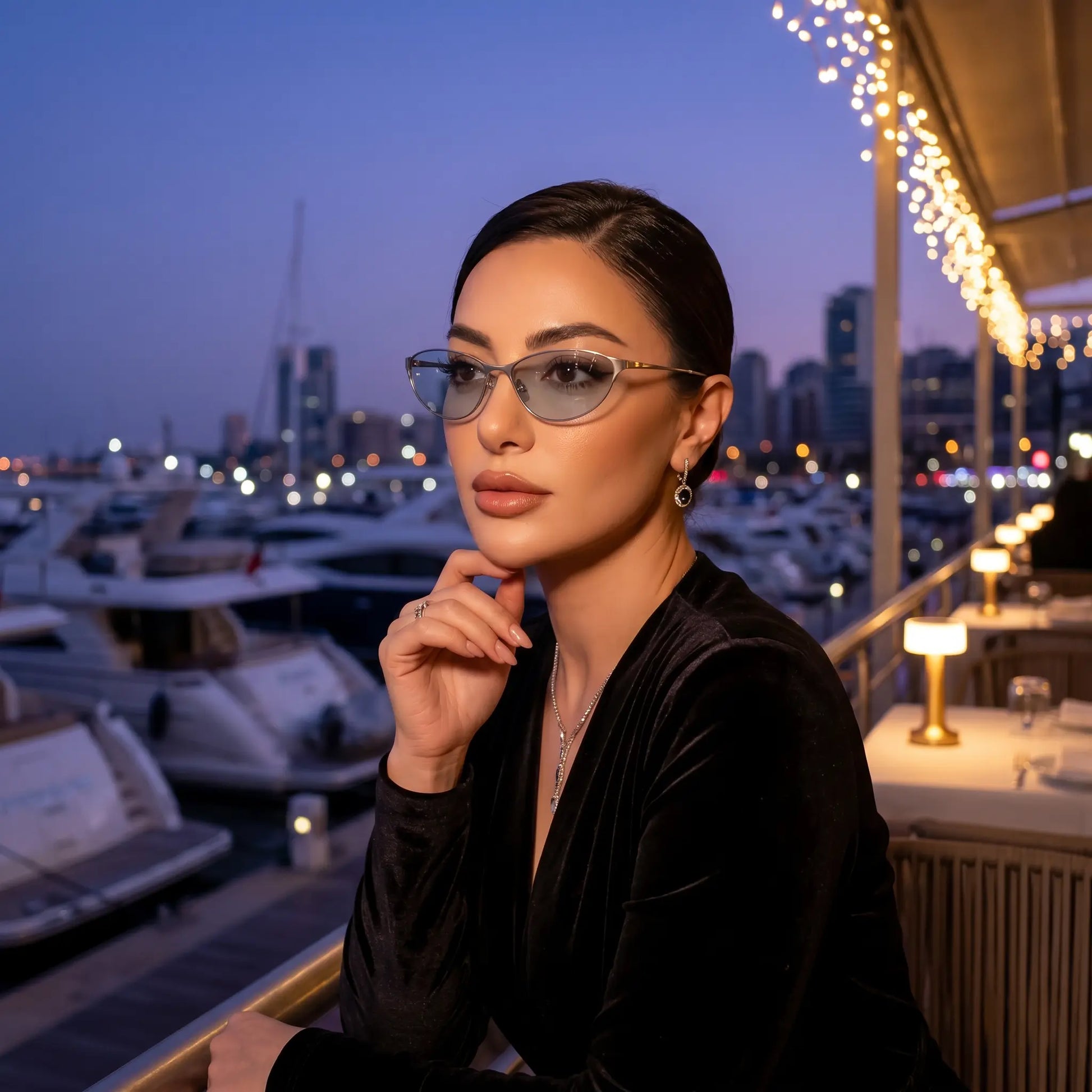 Elegant woman wearing Kivago silver metal cat-eye sunglasses with light blue tinted lenses, posing at a sophisticated outdoor restaurant overlooking a marina at twilight. She wears a black velvet top and delicate jewelry.