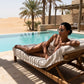 Elegant woman wearing Kivago silver metal cat-eye sunglasses with light blue tinted lenses, relaxing on a lounge chair by a resort pool overlooking a desert landscape.