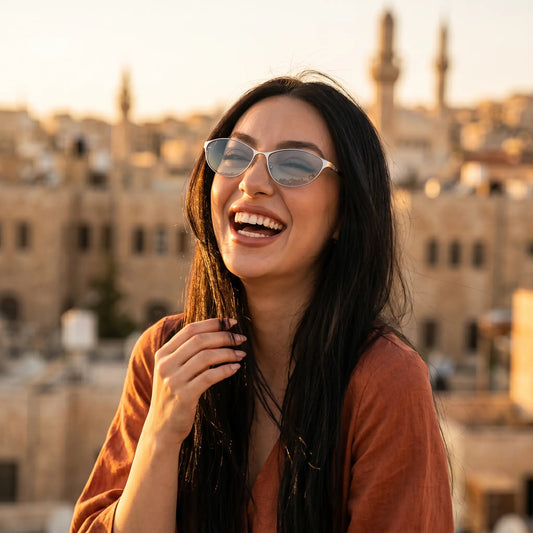 A joyful woman wearing Kivago metal cat-eye sunglasses with silver frames and light blue tinted lenses, smiling brightly outdoors during golden hour with a blurred urban background.