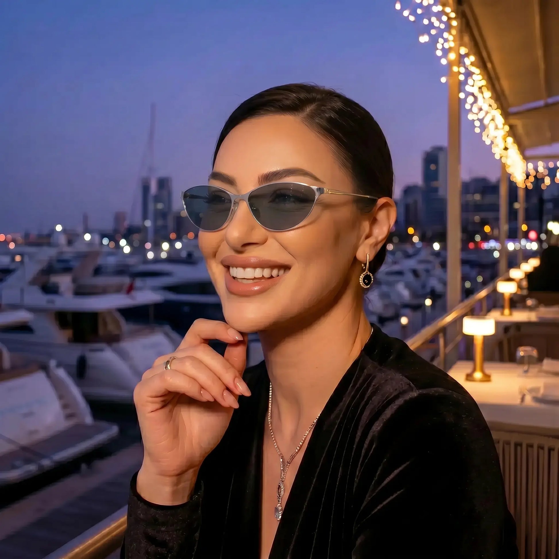 Smiling woman wearing Kivago silver metal cat-eye sunglasses with dark grey tinted lenses, enjoying an elegant evening by a marina.