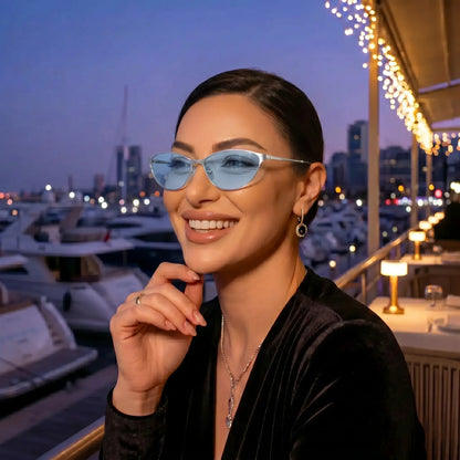 Smiling woman wearing Kivago silver metal cat-eye sunglasses with light blue tinted lenses, enjoying an elegant evening at a marina restaurant.