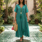 Woman wearing a Lagoon Teal tiered bohemian maxi dress with a V-neck, short sleeves, and crinkled gauze fabric, walking barefoot in a garden, holding a straw tote bag.