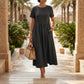 Model wearing a Kivori Midnight Black linen blend midi dress, featuring a round neck, short sleeves, and an empire waist, walking on stone steps in a lush outdoor setting.
