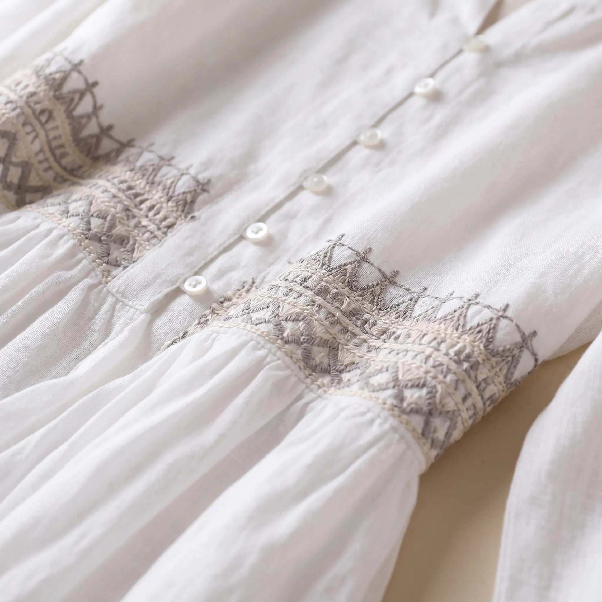 Close-up of the Kivren white bohemian midi dress, showcasing the notched V-neckline, delicate pearl-effect buttons, and intricate geometric embroidery in tan and terracotta threads on the linen-blend empire waist.
