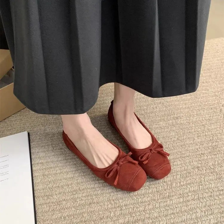 Kivuli Burgundy Wine faux suede ballet flats worn on feet with dark trousers, highlighting the delicate tied bow and square-round toe on a textured rug.