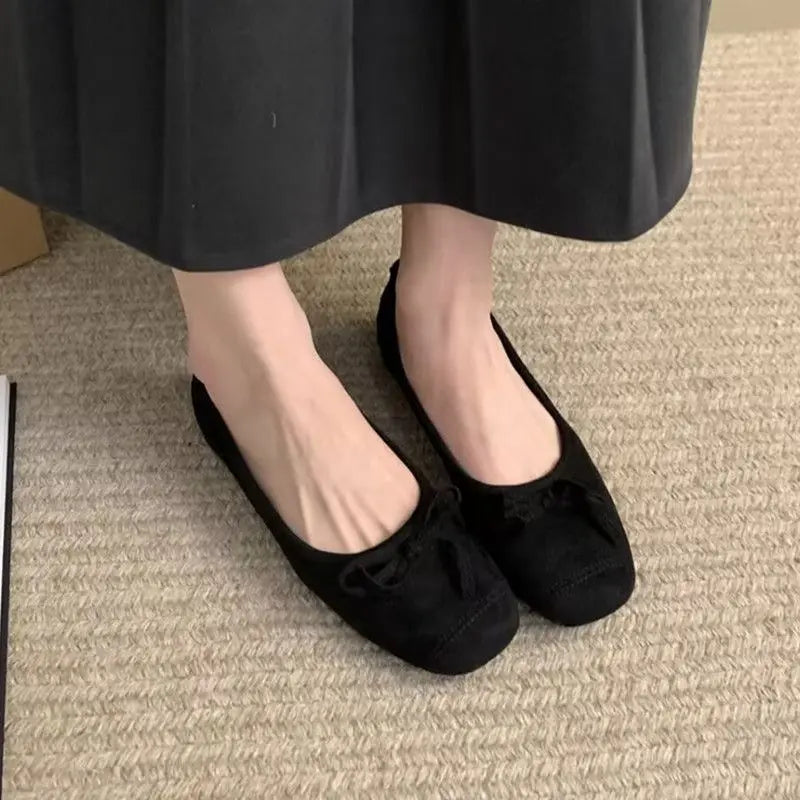 Kivuli Midnight Black faux suede ballet flats, featuring a square-round toe and delicate bow detail, worn on feet with a black skirt on a textured rug.