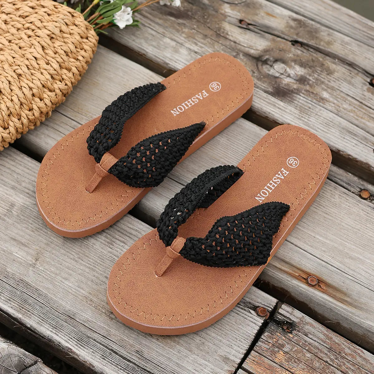 Black woven strap thong flat sandals with brown soles, styled on a rustic wooden surface next to a straw bag.