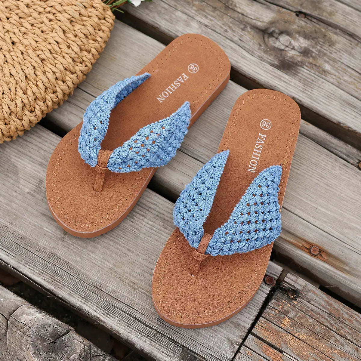 Sky blue woven strap flat sandals with brown insoles and thong toe post, presented on a rustic wooden surface next to a straw bag.