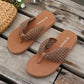 Koreli Bohemian woven flat sandals in brown, shown on a rustic wooden surface with a straw bag and flowers. These comfortable thong sandals feature intricate woven straps and a low-profile design for summer.
