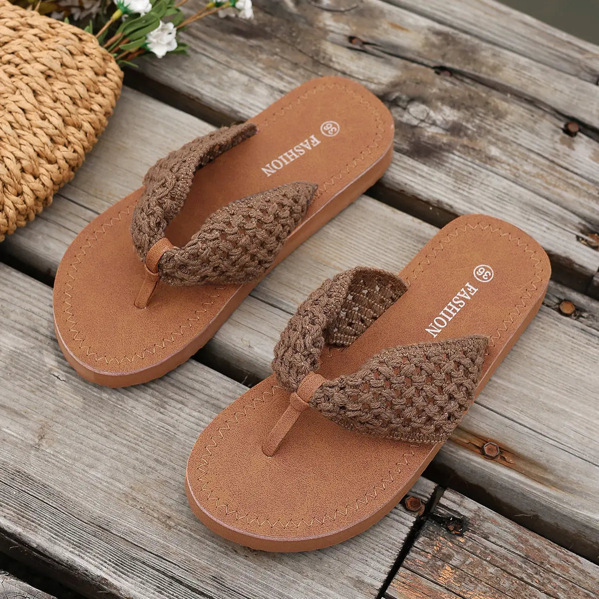 Koreli Bohemian woven flat sandals in brown, shown on a rustic wooden surface with a straw bag and flowers. These comfortable thong sandals feature intricate woven straps and a low-profile design for summer.
