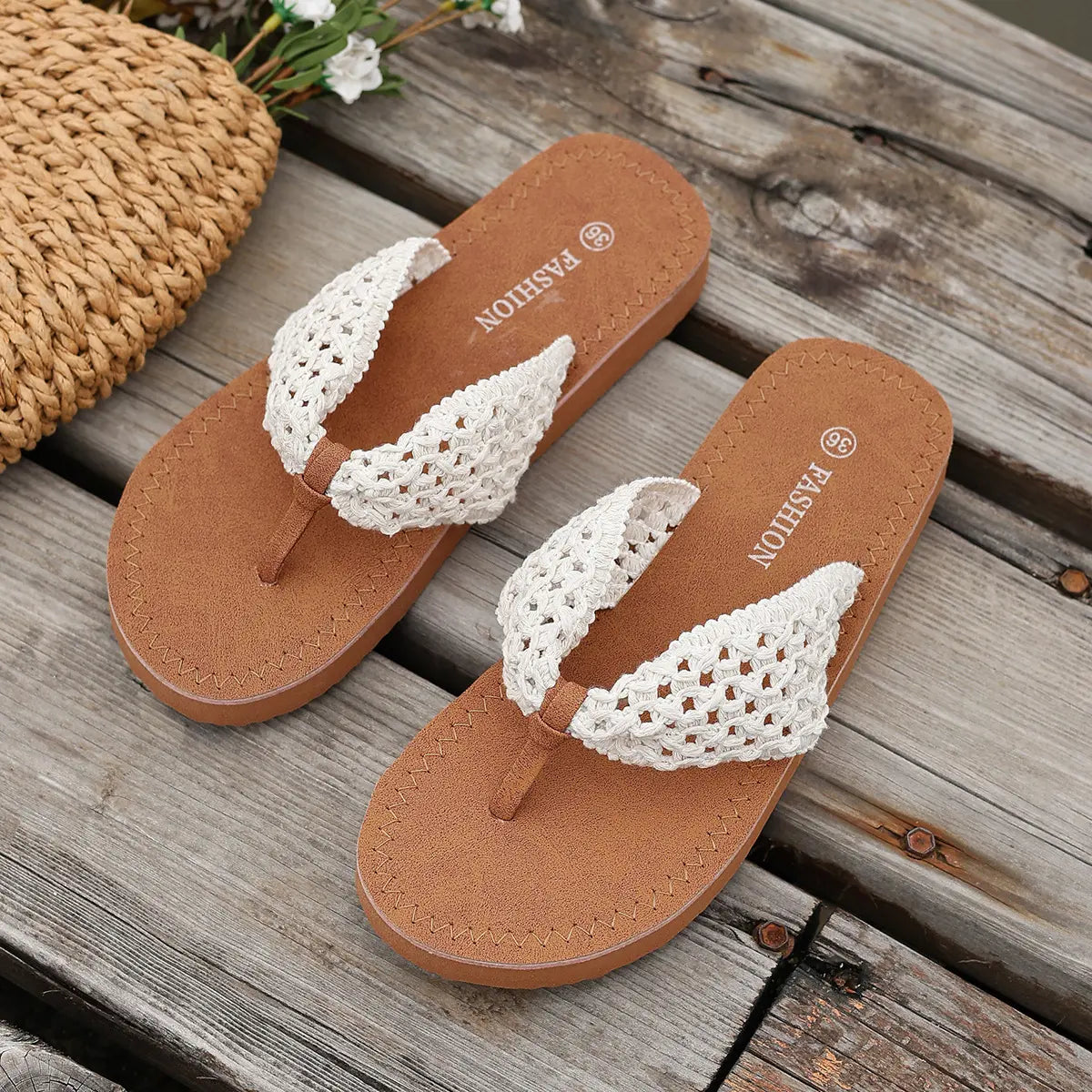Koreli bohemian woven flat sandals in white, displayed on a rustic wooden surface with a straw bag and flowers. The sandals feature intricate woven straps and tan soles.