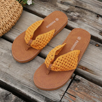 Koreli bohemian woven flat sandals in mustard yellow, shown from a top-down angle on a rustic wooden surface with a straw bag detail. These open-toe thong sandals feature intricate woven straps and a tan sole, perfect for summer.